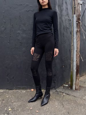 Kvinde, der står mod en tekstureret mørk væg iført sort top med lange ærmer, leggings med blondepaneler og sorte spidse ankelstøvler.