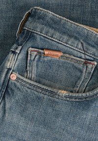 Φωτεινό γαλάζιο τζιν με φθαρμένη υφή, με δερμάτινο έμβλημα που αναγράφει "ARCHIVE" και αντίθετη ραφή στην πίσω τσέπη.