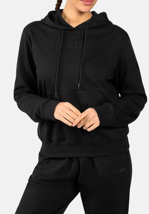 Vrouw draagt een zwarte Reebok hoodie en bijpassende zwarte joggingbroek, handen samengevouwen waarbij een grote gouden ring aan de rechterhand te zien is.