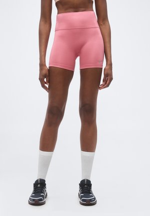 Femeie purtând pantaloni scurți sport roz cu talie înaltă, șosete albe până la gleznă și pantofi negri de alergare stând pe un fundal simplu.