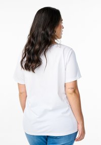 Woman with wavy dark hair wearing a plain white t-shirt and blue jeans, standing facing away with a small tattoo visible on her left arm.