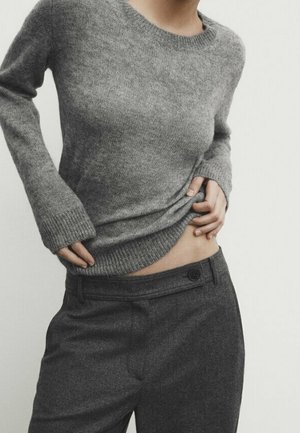 Personne portant un pull en maille gris et un pantalon gris foncé, soulevant légèrement le pull pour révéler une partie de la taille et du bas de l'abdomen.