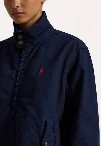 Navyblauwe jas met een hoge kraag, ritssluiting en twee knopen. Heeft een klein rood logo op de linkerborst en zijzakken.