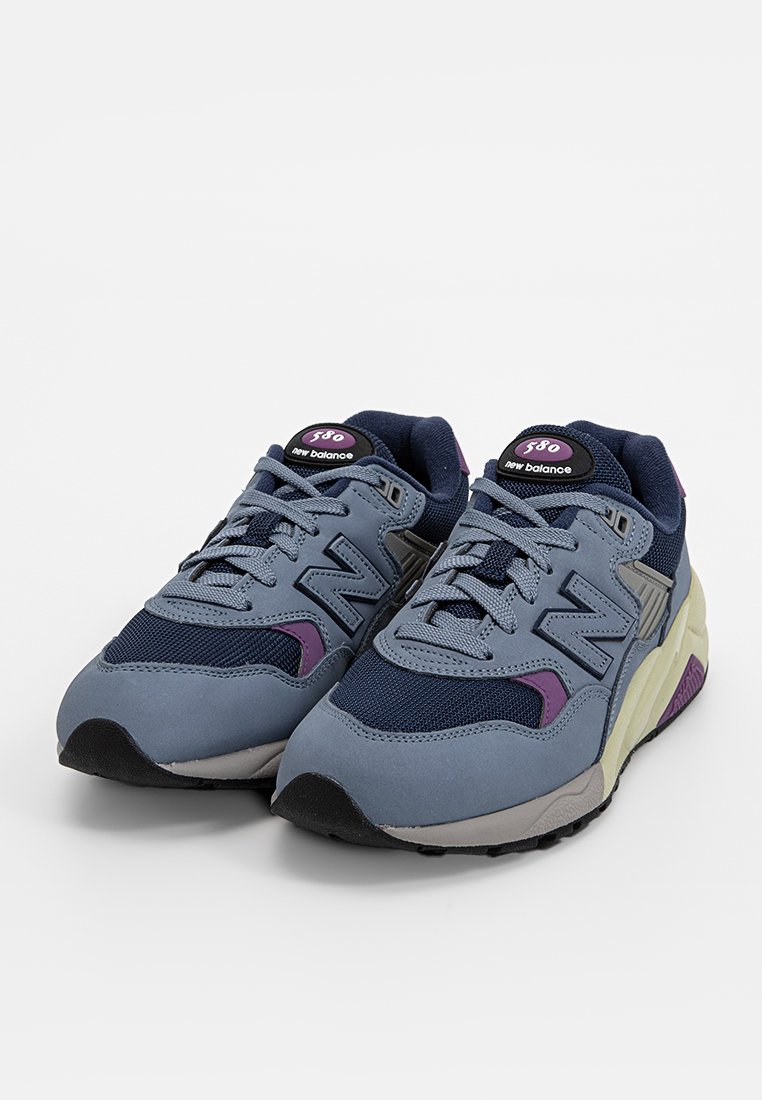 Zapatillas grises y marinas con material de malla y ante, con un acento púrpura, cordones prominentes y el logo de "New Balance" en la lengüeta.