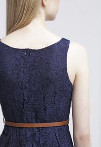 Marineblauw kanten jurk met een aangesloten bovenstuk en een bruine leren riem om de taille. Toont bloemenpatroon en een scoop rugdecolleté.