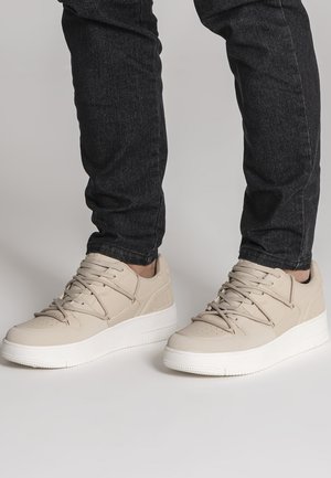 Beige flache Sneaker mit weißen Sohlen, getragen zu dunklen Skinny Jeans, dargestellt von der Mitte der Wade abwärts vor neutralem Hintergrund.