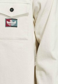 Camisa de pana color crema con un bolsillo en el pecho que lleva un parche con un logo colorido de un pájaro. La tela presenta un patrón acanalado texturizado.