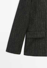 Blazer gris foncé en tissu texturé subtil, avec une poche poitrine unique et un détail de manche ajusté. Sans fermetures visibles ni ornements.