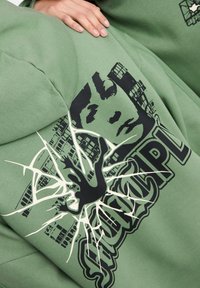 Sudadera verde con un gran gráfico negro que presenta una cara, una mano y elementos de diseño desgastados. El material parece suave y texturizado.