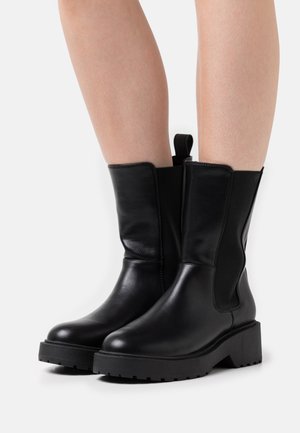 Black ankle boots made of leather with elastic side panels and a pull tab. Features a chunky black sole with a treaded pattern.
