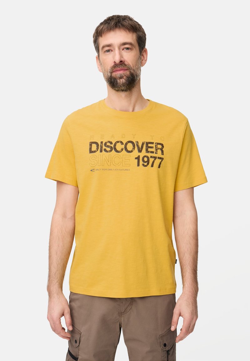 Mann mit Bart, der ein gelbes T-Shirt mit dem Aufdruck "DISCOVER SINCE 1977" und braune Cargo-Hosen trägt, steht vor weißem Hintergrund.