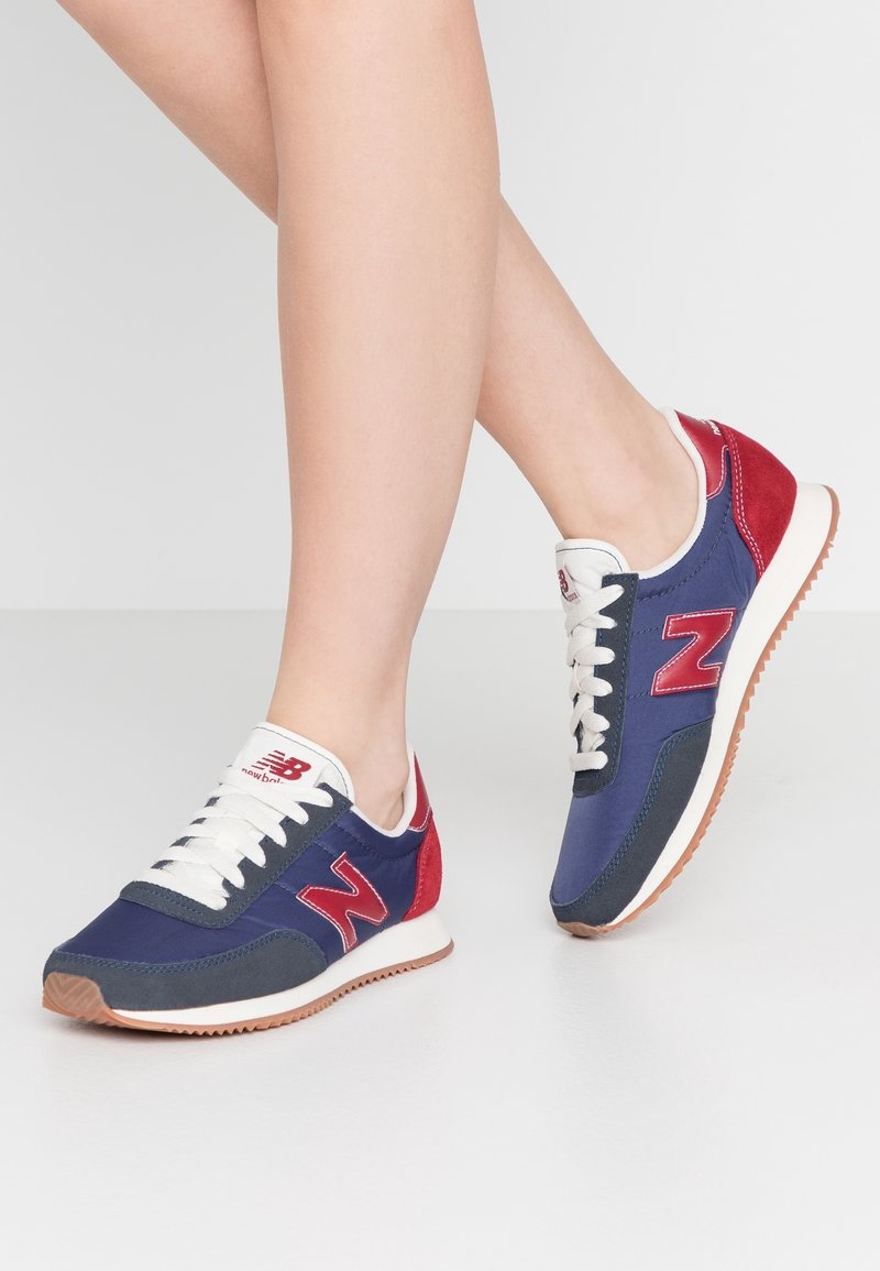 Chaussures de sport bleu marine et rouge avec une tige en tissu texturé, des lacets blancs et une semelle en caoutchouc marron, arborant un logo "N" rouge proéminent.