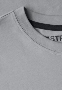 Světle šedé tričko s kulatým výstřihem a tmavě šedým lemem. Tkanina je hladká s jemnou strukturou a čistými stehy.