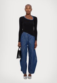 Maglietta nera a maniche lunghe con dettagli di cucitura blu, abbinata a jeans blu a gamba larga con cuciture visibili. Il modello tiene in mano una piccola borsa nera.