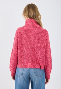Maglione lavorato a maglia di un vivace rosa con colletto alto e vestibilità ampia. Presenta un tessuto testurizzato con polsini e vita a coste. Abbinato a jeans blu.