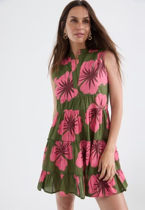 Mujer con vestido verde sin mangas con gran estampado floral rosa y pendientes de aro dorados sobre un fondo claro liso.