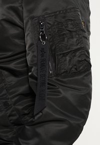 Schwarzer, gesteppter Jackenärmel mit strukturierter Oberfläche, ausgestattet mit einer Reißverschlusstasche und einem auffälligen Stoffetikett mit schwarzer Schrift.