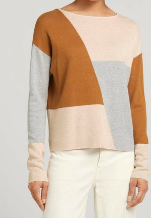 Femme portant un pull à manches longues à blocs de couleurs en beige, rouille et gris, associé à un pantalon blanc, vue du cou jusqu'aux hanches.