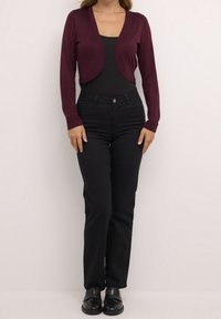 Bordeaux geknipte cardigan met lange mouwen, zachte textuur en open voorkant, gecombineerd met zwarte high-waisted jeans en zwarte schoenen.