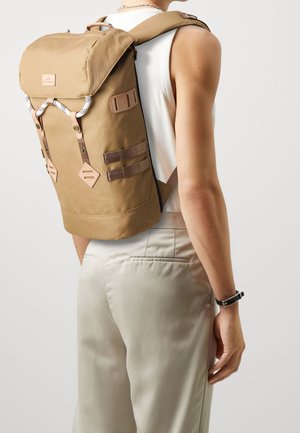 Sac à dos en toile marron avec un design à rabat enroulable. Il est doté de bretelles beige, d'un accent en tissu sur le rabat et de détails en métal à l'extérieur.