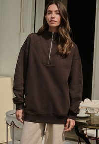 Sweat-shirt marron à fermeture éclair avec col montant, doté d'une grande poche avant et de poignets côtelés, porté sur un pantalon clair et texturé.
