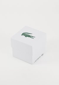 Boîte blanche avec une finition texturée et un logo de crocodile vert en haut. Le côté présente le texte imprimé "LACOSTE." Forme carrée.