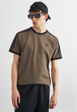 Ung mand iført en brun Adidas t-shirt med sorte striber, sort kasket bagvendt, oversize sorte solbriller og sorte bukser, stående mod en hvid baggrund.