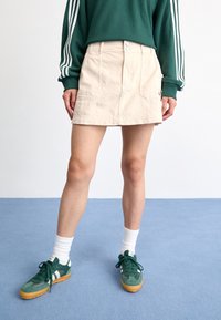 Beige spijkerrok met voorkruiden en twee knoopsluitingen, gecombineerd met een groene sweatshirt en groene sneakers met witte accenten.