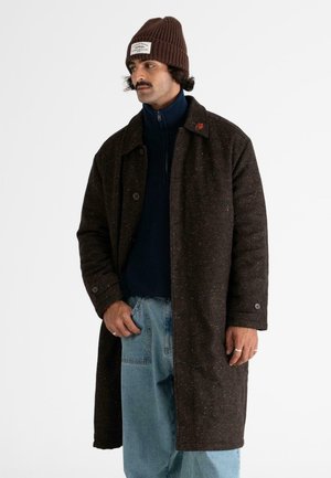 Braun gesprenkelter Wollmantel mit großen Knöpfen, getragen über einem marineblauen Pullover und hellblauen Jeans. Die Accessoires umfassen eine braune Strickmütze.