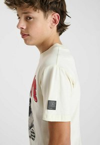 T-shirt crème à manches courtes avec un imprimé graphique et un patch logo sur la manche. Matériau en coton doux avec une coupe décontractée.