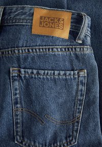 Traperice tamnoplave boje s kožnom oznakom u boji pijeska s natpisom "JACK & JONES", s žutim šavovima i prepoznatljivim dizajnom stražnjeg džepa.