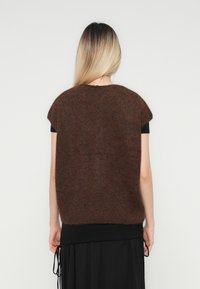 Frau mit langen blonden Haaren, die einen braunen, fuffy ärmellosen Pullover über einem schwarzen Kleid trägt, mit dem Rücken zu einem schlichten weißen Hintergrund gewandt.
