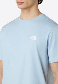 Camiseta de algodón azul claro con cuello redondo, mangas cortas y un pequeño logo blanco en el lado izquierdo del pecho, con una textura suave.