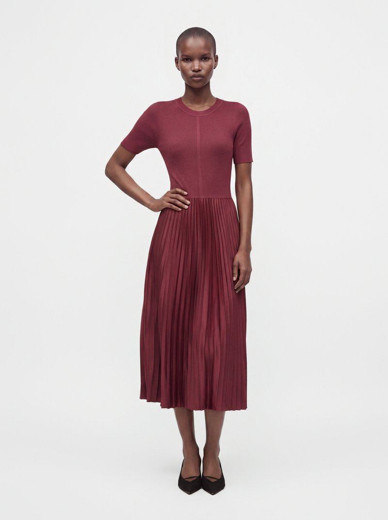 Femme debout, main sur la hanche, portant un top ajusté à manches courtes et une robe jupe midi plissée rouge foncé avec des chaussures noires pointues.