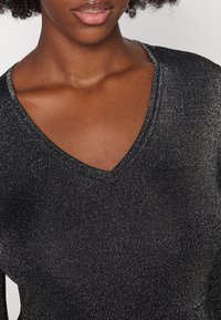 Haut en maille noir scintillant avec un décolleté en V profond. Le matériau a un éclat métallique et une texture lisse, mettant en valeur le design élégant.