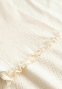 Cremefarbener Stoff mit strukturierten Streifen und kleinen Lochmustern, der an einer Kante mit einem Rüschensaum verziert ist.
