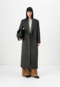Femme avec des cheveux bruns lisses portant un long manteau gris foncé à double boutonnage, un col roulé rayé, un pantalon large marron, tenant une grande pochette noire.