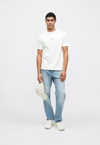 Valge puuvillane t-särk väikese tekstilogoga, hele sinised teksad, käes beež nokamüts ja jalas valged tossud. Lihtne, vabaaja stiilis riietus.