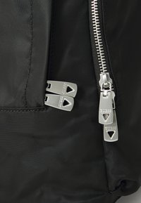 Sac à dos en nylon noir avec des fermetures éclair argentées, des tirettes à double zip arborant le logo "GUESS" et un détail de surface texturé.