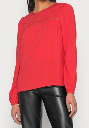 Red long-sleeve blouse with lace detail across the chest, worn with black leather pants, shown from shoulders to hips.