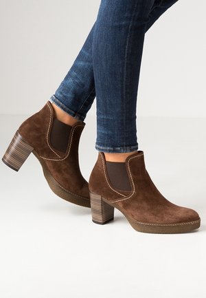 Brune ruskind ankle støvler med stablede blokhæle båret med opkørte blå skinny jeans på en lys baggrund.