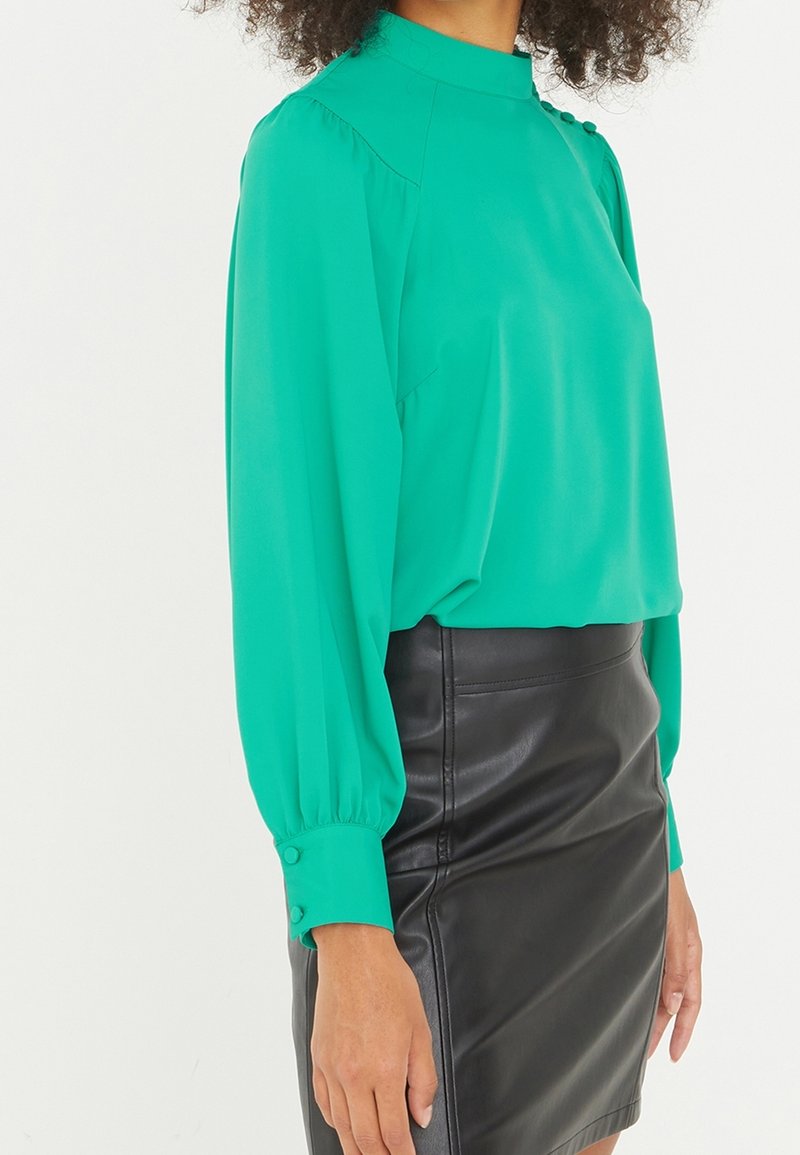 Green blouse with a high neck, long puff sleeves, and button details on cuffs, paired with a black leather mini skirt.