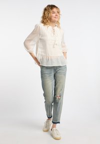 Bílá blouse s puntíky, s peplum lemem, zavázaným výstřihem a dlouhými vypouklými rukávy, v kombinaci se světlými modrými roztrhanými džíny a bílými teniskami.