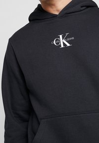 Felpa nera con cappuccio realizzata in tessuto testurizzato, dotata di una tasca frontale a marsupio e di un logo CK bianco con "CALVIN KLEIN JEANS" sottostante.