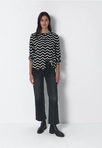 Donna in piedi contro un muro bianco, che indossa una blusa a maniche lunghe con motivo zigzag in bianco e nero, pantaloni neri cropped e stivaletti neri.