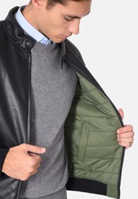 Giacca di pelle nera con interno trapuntato verde, dotata di chiusura a scatto, collo alto e polsini a coste. Maglione grigio sotto.