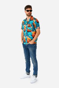 Camicetta a maniche corte con colletto in una vivace stampa tropicale, con sfondo blu e elementi arancioni e rossi. Indossata con jeans blu e sneakers bianche.