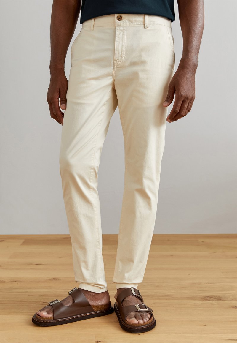 Chinos en coton beige clair avec une coupe ajustée, dotés d'une fermeture à bouton à l'avant et de poches latérales, associés à des sandales en cuir marron.