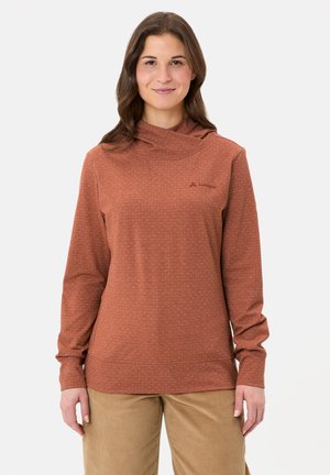 Frau trägt einen rostfarbenen Pullover mit langen Ärmeln und dezenten Punkten sowie eine beigen Hose, steht und blickt nach vorne vor weißem Hintergrund.