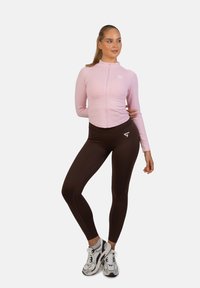 Lys rosa lynlåset langærmet top parret med brune leggings; begge lavet af strækbart stof. Bemærkelsesværdige hvide logo-accenter på hvert stykke.